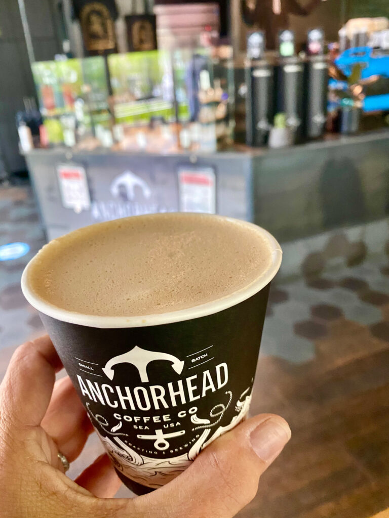 A close-up of a hand holding a cup of coffee in a black Anchorhead Coffee Co. cup, with a blurred cafe counter in the background. The scene suggests a cozy coffee shop environment.