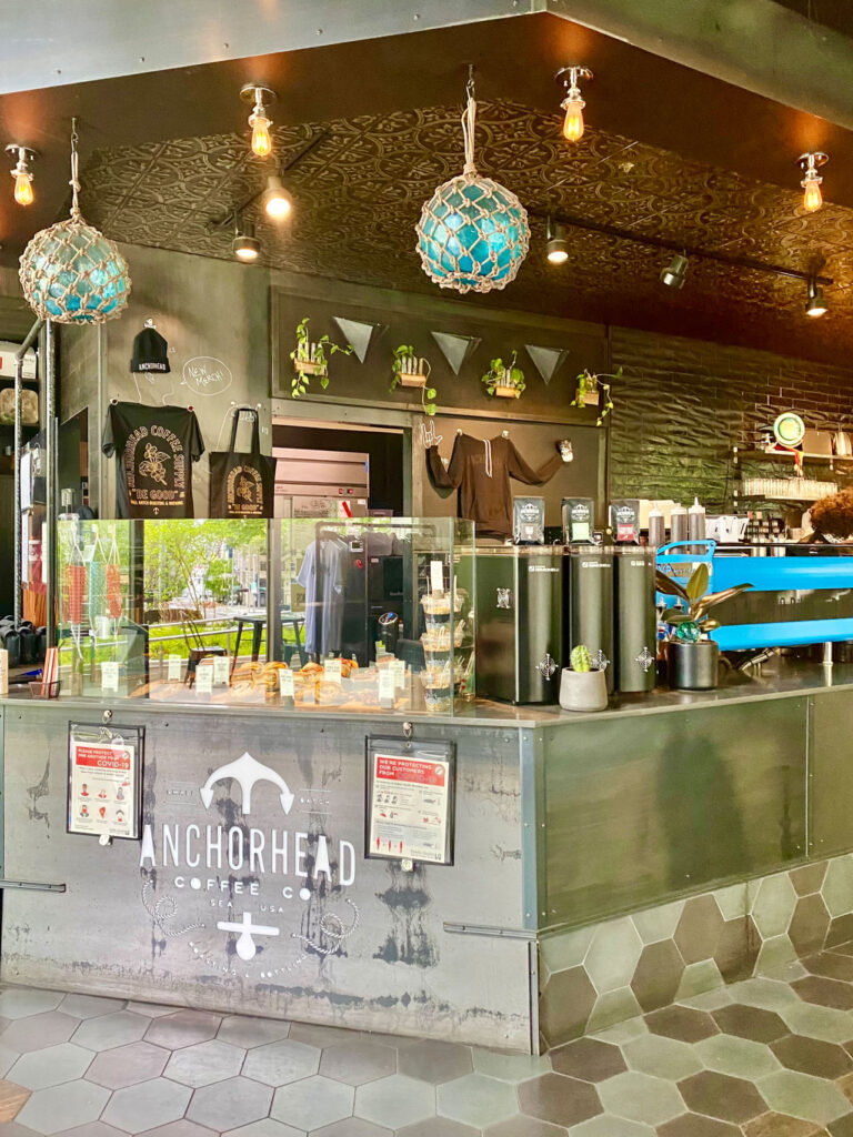 A cozy coffee shop interior featuring a counter with a glass display of pastries. The decor includes hanging lights, potted plants, and stylish overhead details. Prominent branding on the metal counter reads "Anchorhead Coffee Co.