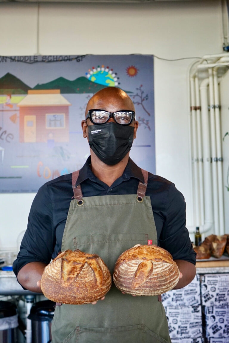 A person wearing glasses and a mask stands indoors, holding two loaves of artisan bread. They are dressed in a black shirt and olive green apron, with a colorful poster on the wall behind them.