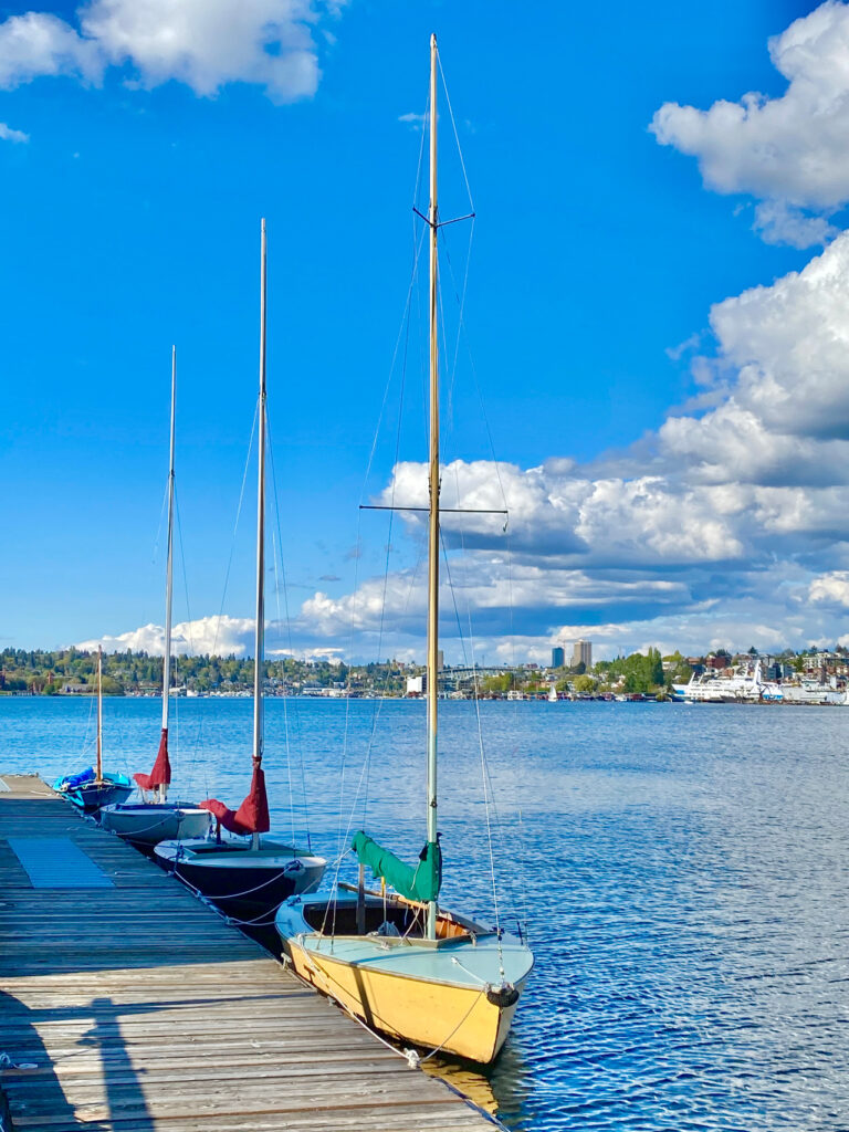 Three sailboats docked at a wooden pier on a clear blue lake under a sky dotted with fluffy clouds. The boats have tall masts, and the background shows a shoreline with buildings and greenery.