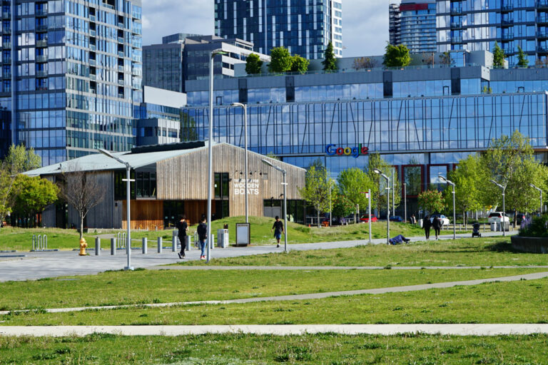 A modern urban park with grassy areas and pathways is in the foreground. People are walking and enjoying the sunny day. Behind the park are glass buildings, including one with a prominent Google logo. Trees and benches are scattered throughout.
