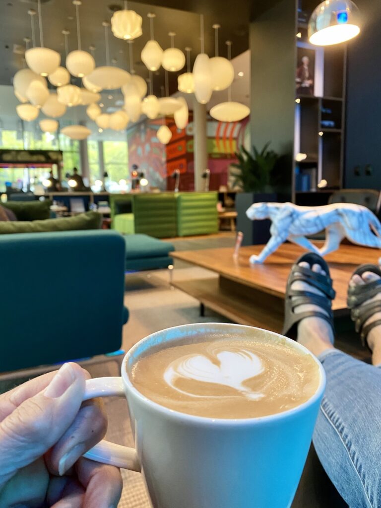 A cozy cafe with modern decor, featuring hanging lights and dark furniture. A person holds a mug of latte with latte art, while relaxing with sandals on a table. Colorful mural in the background.