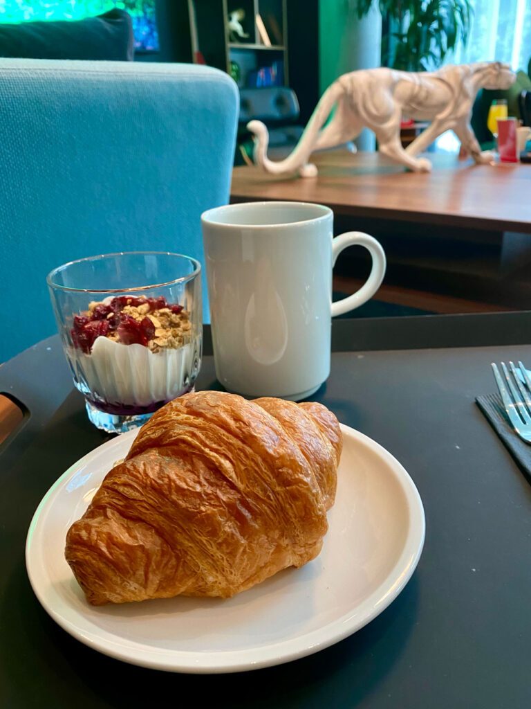 A croissant on a white plate, a glass of yogurt with granola and berries, and a white mug sit on a dark table. In the background, there's a white tiger sculpture on a wooden table and a blue chair.