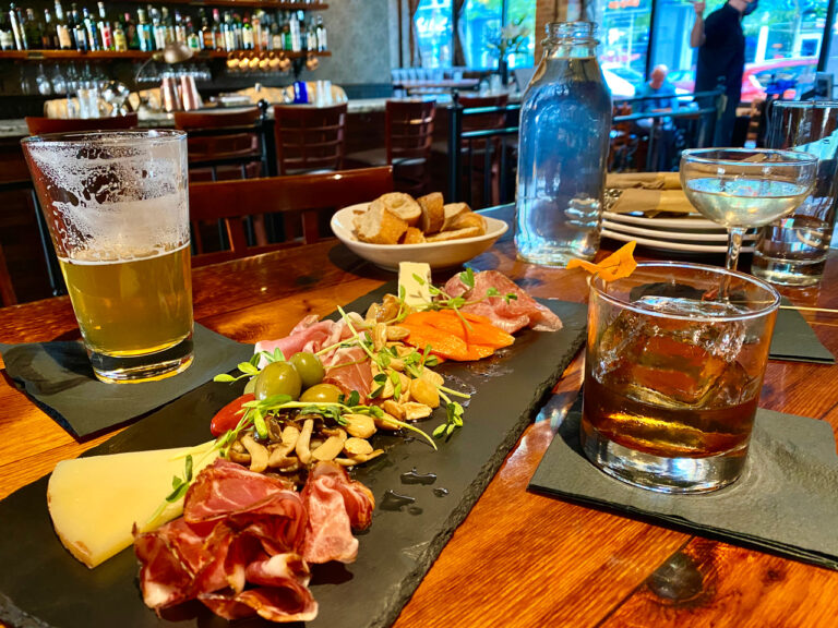 A slate platter with cured meats, cheese, olives, and nuts is on a wooden table. Nearby, there's a pint of beer, a glass of whiskey, a cocktail, a bottle of water, and a bowl of bread. The setting is a cozy bar or restaurant.