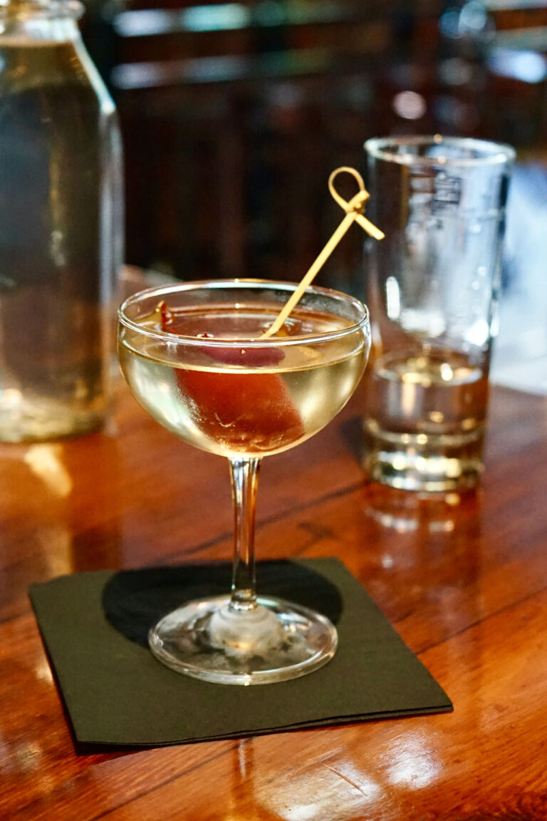 A cocktail in a coupe glass garnished with a skewer and an olive, placed on a black napkin on a wooden table. In the background, there's a glass and a water bottle, with a blurred bar setting.