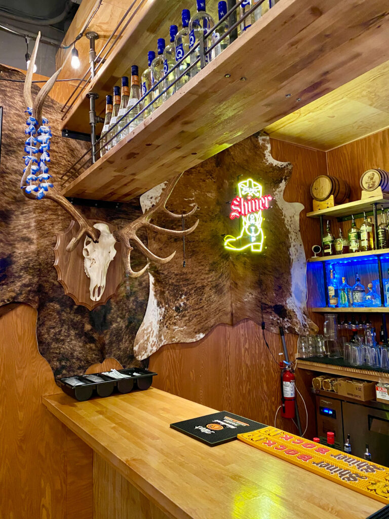 A rustic bar interior featuring a wooden counter and shelves lined with liquor bottles. A neon Shiner Beers sign and antlers mounted on an animal hide are on the wall. A fire extinguisher and various bar tools are visible.