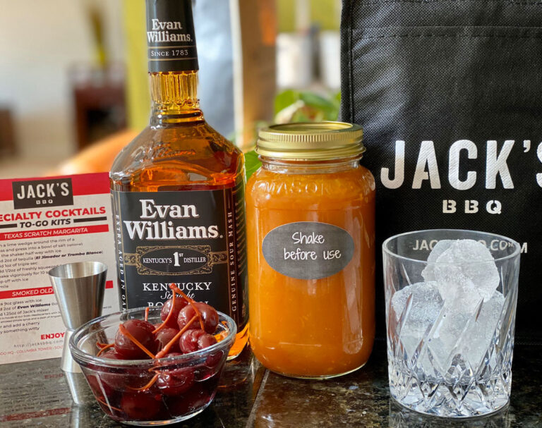 A bottle of Evan Williams bourbon, a jar labeled "Shake before use" containing an orange liquid, a bowl of cherries, a glass with ice cubes, a jigger, and a Jack's BBQ bag are arranged on a countertop.