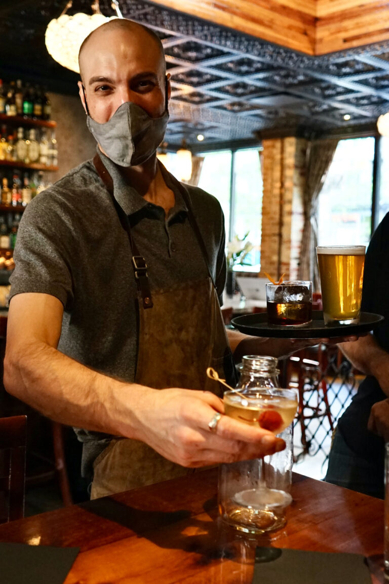 A waiter wearing a gray shirt and apron serves drinks in a dimly lit pub. He holds a tray with a beer and another drink while offering a jar with a cocktail to a customer. Shelves with bottles are visible in the background.
