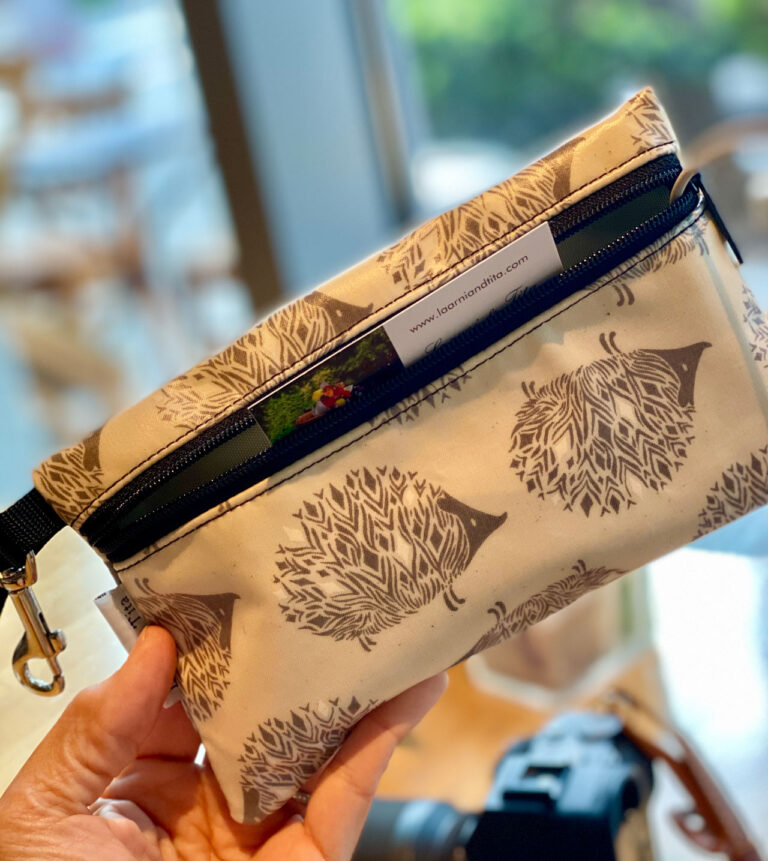 A person holds a beige pouch with a zipper, adorned with hedgehog illustrations. The pouch has a strap with a metallic clasp. A website URL is visible on a small white label attached to the pouch.