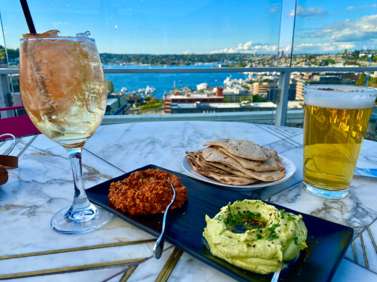 A table on a rooftop with a view of the city and water. On the table are a glass of white wine with ice, a pint of beer, a plate of flatbreads, and a black dish with red dip and green dip, garnished with herbs.