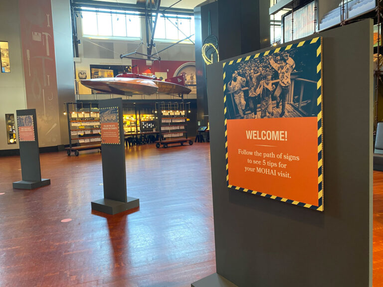 Museum interior with wooden floors and high ceilings. A welcome sign with instructions is in the foreground. A suspended, curved object and display cases are in the background. Exhibition lighting highlights the displays.
