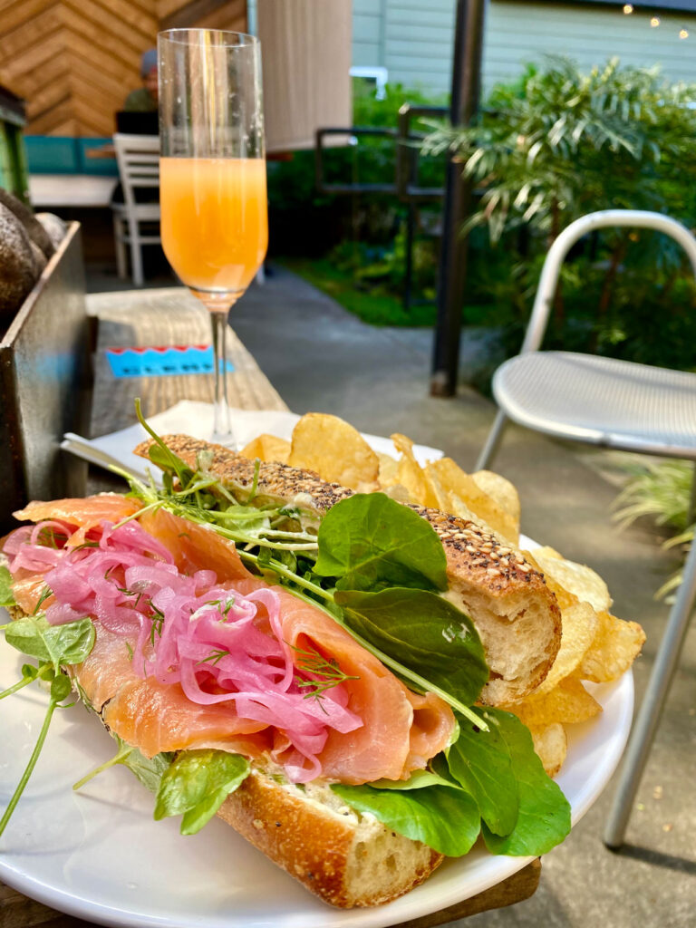 A gourmet sandwich with smoked salmon, pickled onions, and greens on a seeded baguette is served with a side of potato chips on a white plate. A tall glass of orange juice is in the background beside a metal chair in a garden setting.