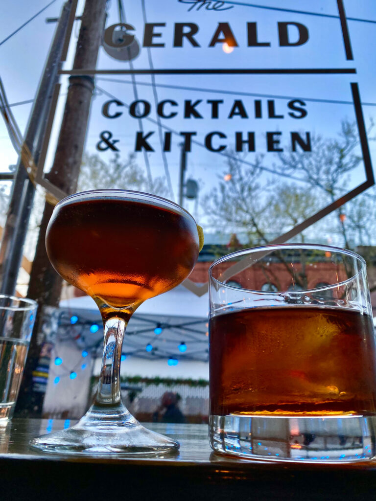 Two cocktails on a wooden table in front of a window displaying the words "COCKTAILS & KITCHEN" with outdoor string lights and trees in the background. The setting appears relaxed and inviting.