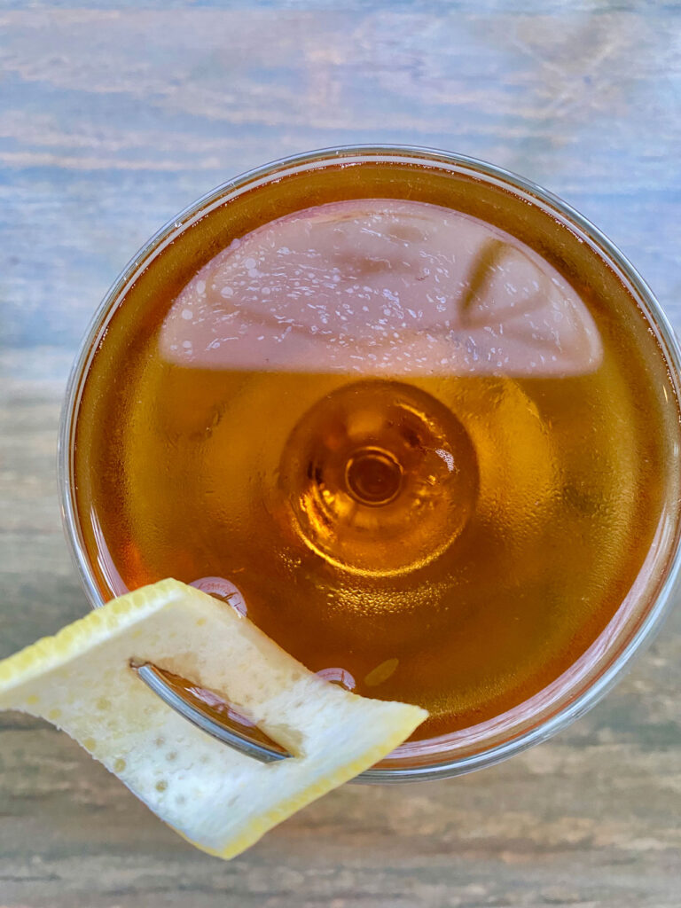 Overhead view of a brown cocktail in a glass with a lemon twist garnish on the rim. The background is a wooden table.