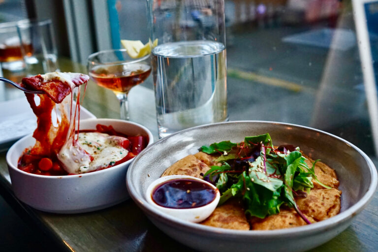 A table setting with a plate of pizza topped with greens and a small dish of sauce. Nearby is a bowl of food being forked, a glass of water, and a cocktail, against a blurred window background.