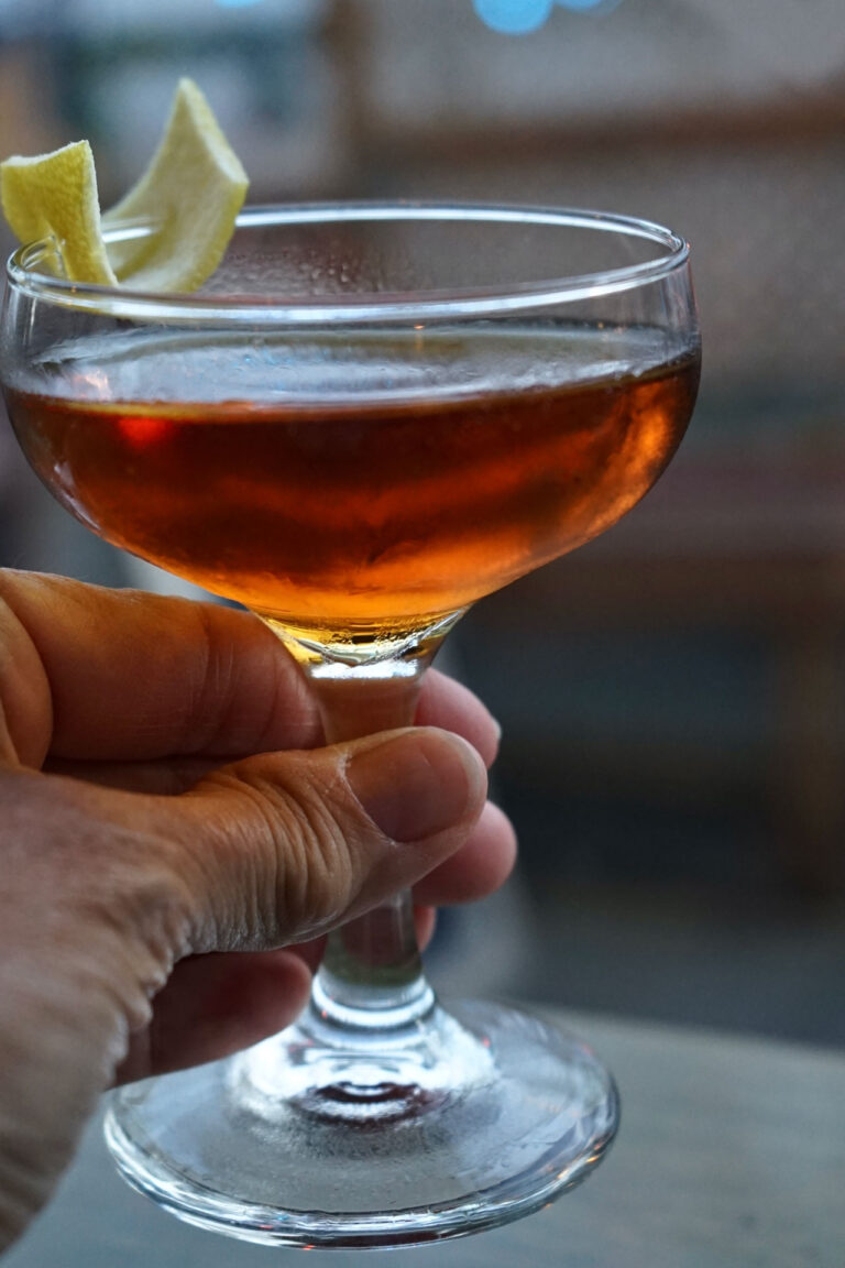 A hand holding a coupe glass filled with an amber-colored cocktail, garnished with a lemon twist. The background is softly blurred, suggesting a cozy indoor setting.