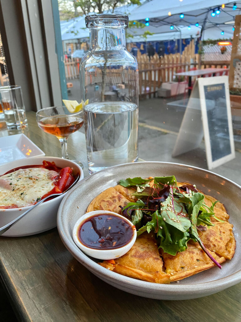 A bowl of baked dish with red sauce and cheese beside a plate of savory pancakes topped with greens and a small bowl of brown sauce. A glass bottle of water and a cocktail are on the wooden table by a street-view window.