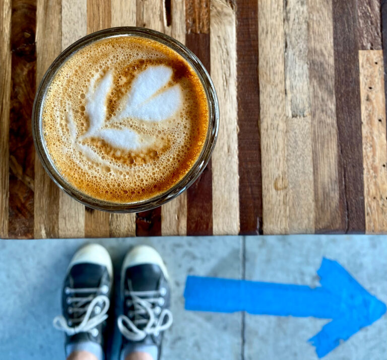 A glass of coffee with latte art on a wooden table. Below, a pair of sneakers stands on a concrete floor, pointing towards a blue arrow made of tape indicating a direction.