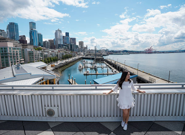 A person in a white dress stands on a balcony overlooking a city skyline with tall buildings and a marina. The sky is partly cloudy, and the water is calm, reflecting the blue of the sky.