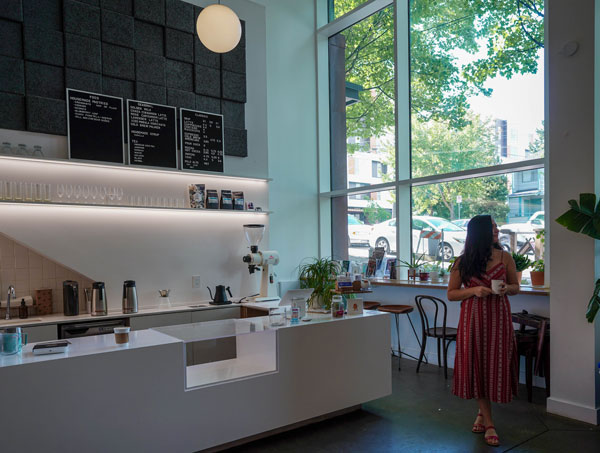 A woman in a red dress stands inside a modern café, holding a cup. The café features a sleek white counter, menu boards on a dark wall, plants, and large windows showing trees and parked cars outside.
