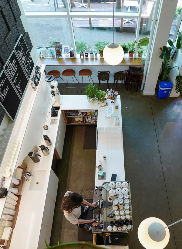 Aerial view of a modern coffee shop with a barista preparing drinks. The space features white countertops, wooden chairs, a large menu board, and greenery. Large windows provide natural light, and there are indoor plants decorating the interior.