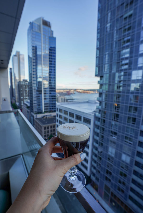 A hand holding a glass of coffee with a frothy top near a balcony railing. Tall city skyscrapers and a river in the background under a clear blue sky.