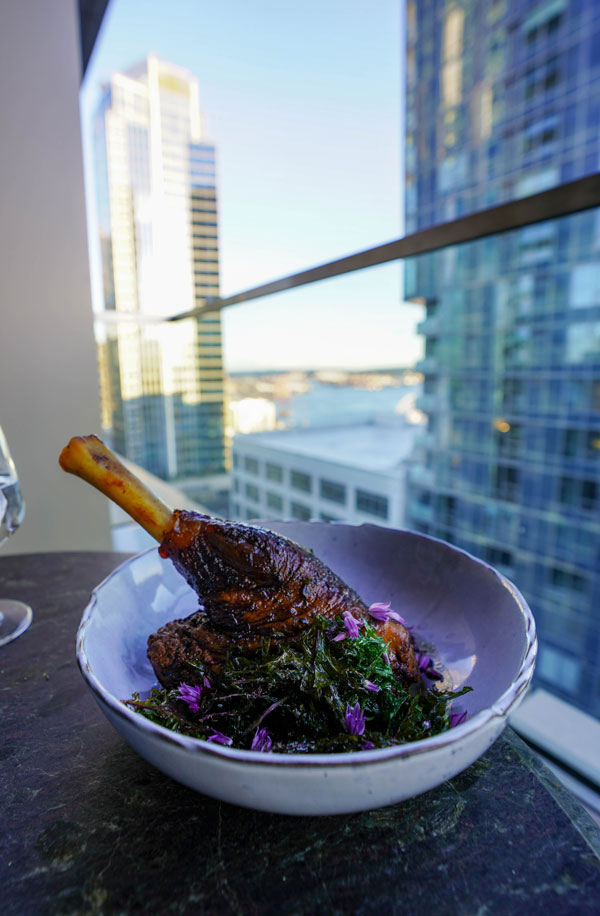 A bowl with a cooked lamb shank garnished with herbs and small purple flowers sits on a table. The background features tall city buildings viewed from a balcony on a sunny day.