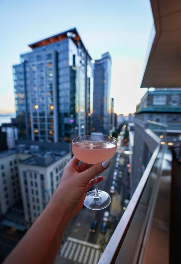 A person holds a cocktail in a stemmed glass, with a cityscape view in the background. Tall buildings line a bustling street as dusk falls, creating a vibrant urban atmosphere.