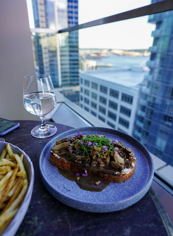 A plate of toast topped with mushrooms, greens, and flowers on a table, accompanied by fries and a glass of water. The setting includes a high-rise view of city buildings and a body of water in the background.