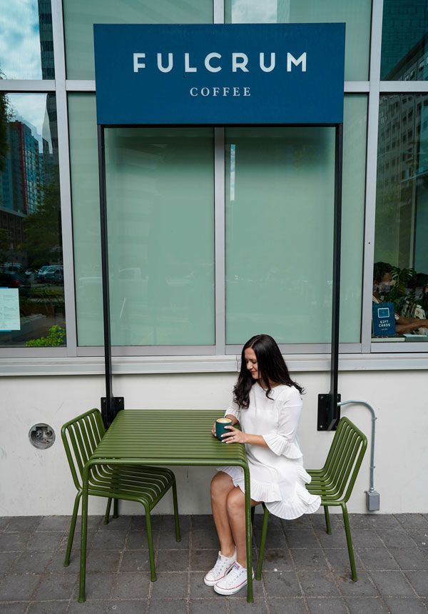 A person in a white dress sits at a green table outside Fulcrum Coffee, holding a blue cup. The café's sign is visible above. Urban buildings and trees are reflected in the window behind.