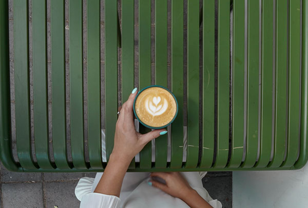 A person with light blue nail polish holds a cup of coffee with latte art, featuring a heart design. The person is sitting on a green slatted bench, and their white sleeve is partially visible.
