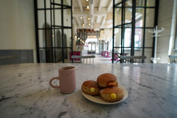 A marble table in a spacious cafe displays a pink mug filled with a beverage next to a white plate of pastries. The background features glass doors, high ceilings, and scattered seating.