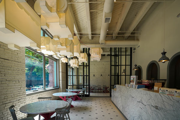 Modern café interior featuring high ceilings with exposed beams and geometric light fixtures. Large windows provide natural light, illuminating marble-topped tables and a marble counter. The space is decorated in a minimalist style.