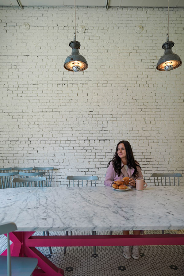 A woman with long dark hair sits alone at a large white marble table, holding a mug and looking to the side. There are pastries in front of her. The background features a white brick wall and hanging industrial lights.