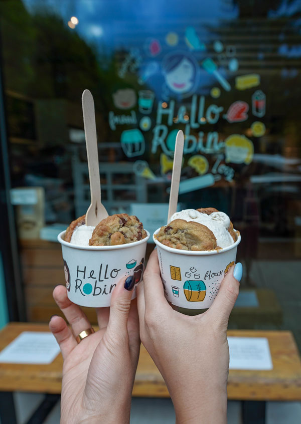 Two hands holding cups of ice cream with wooden spoons in front of a store window decorated with colorful images and the text "Hello Robin." The cups have playful designs, and cookies are visible on top of the ice cream.