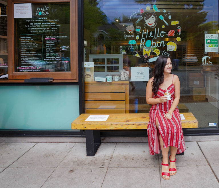 A woman in a red striped dress sits on a wooden bench outside an ice cream shop. She is holding a cup of ice cream and smiling, with the shop's colorful window display in the background.