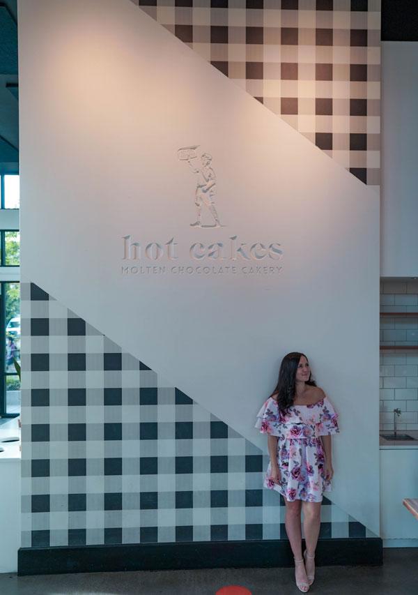 A woman in a floral dress stands against a checkered wall at a location with the name "hot cakes molten chocolate cakery" displayed.