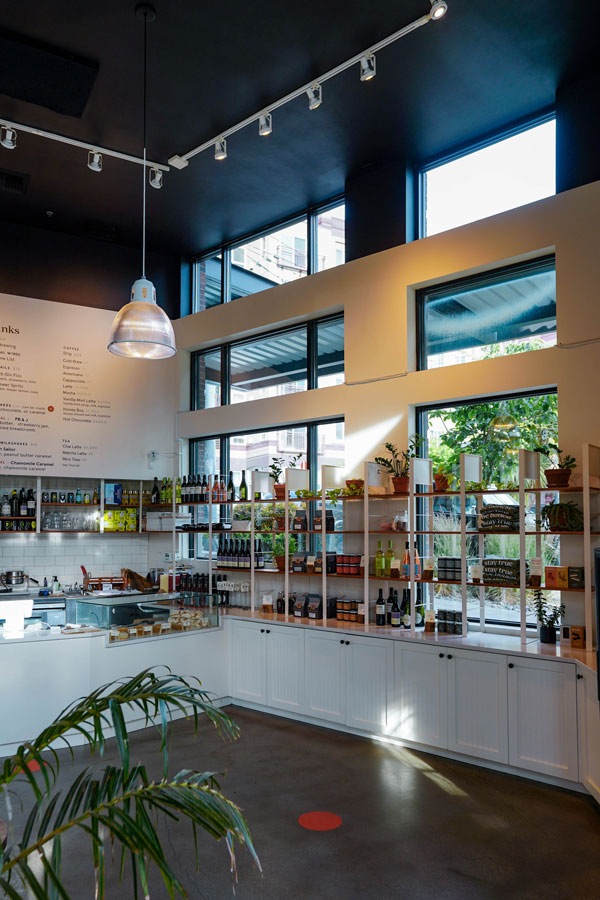 A modern indoor café with high ceilings, large windows, and shelves displaying bottles and snacks. A hanging lamp illuminates the counter area. Plants add a touch of greenery to the space.