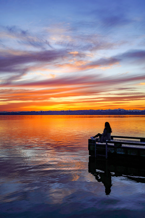 A person sits peacefully on a dock, gazing at a stunning sunset over a calm lake. The sky is filled with vibrant hues of orange, purple, and blue, reflecting beautifully on the water's surface.