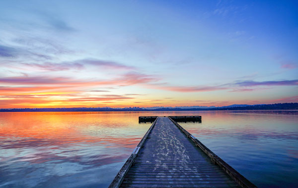 A wooden pier stretches out into a calm lake under a vibrant sunset. The sky displays a blend of orange, pink, and purple hues reflected on the water's surface, creating a serene and picturesque scene.