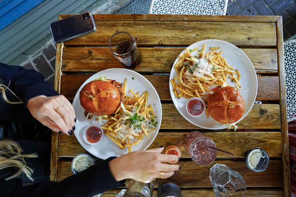 Two people sitting at a wooden table with burgers and fries on white plates, accompanied by dipping sauces. A smartphone and two glasses of soda are placed on the table. They are reaching for the food and drinks.