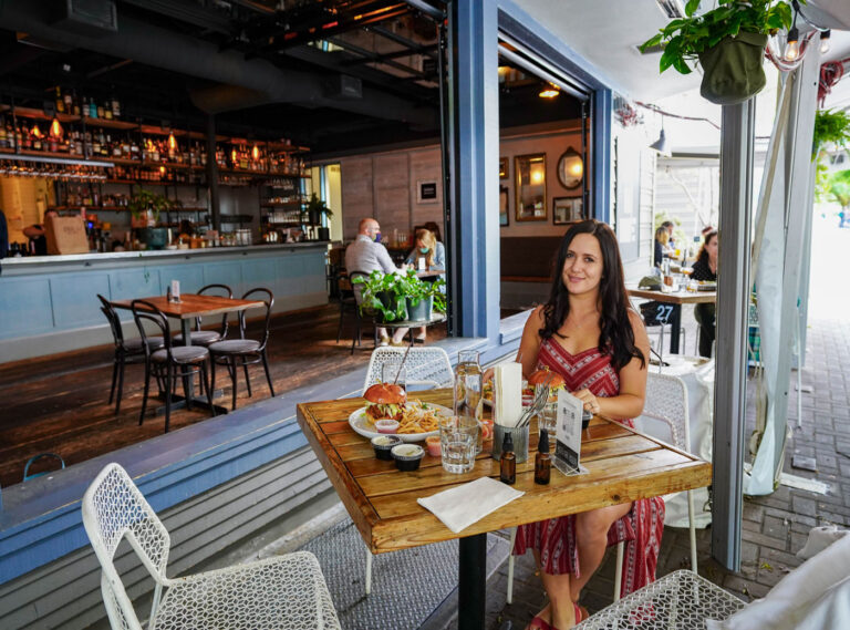 A woman is seated at a wooden table in an outdoor restaurant. She is smiling, with food and drinks in front of her. The background shows an indoor bar area with additional seating and plants hanging above.