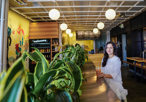 Woman in white dress sitting at a cafe counter holding a drink. The interior features plants on the counter, round hanging lights, wall art, and a menu board in the background.