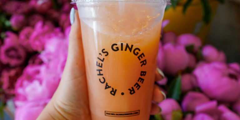A hand holding a plastic cup filled with pink ginger beer. The cup has the label "Rachel's Ginger Beer." In the background, there are vibrant pink flowers, slightly out of focus.