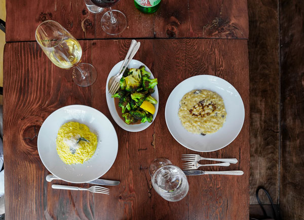 Two plates of pasta and risotto are arranged on a wooden table, accompanied by a dish of vegetables and drinks. The setting includes wine glasses and water, suggesting a cozy dining experience.