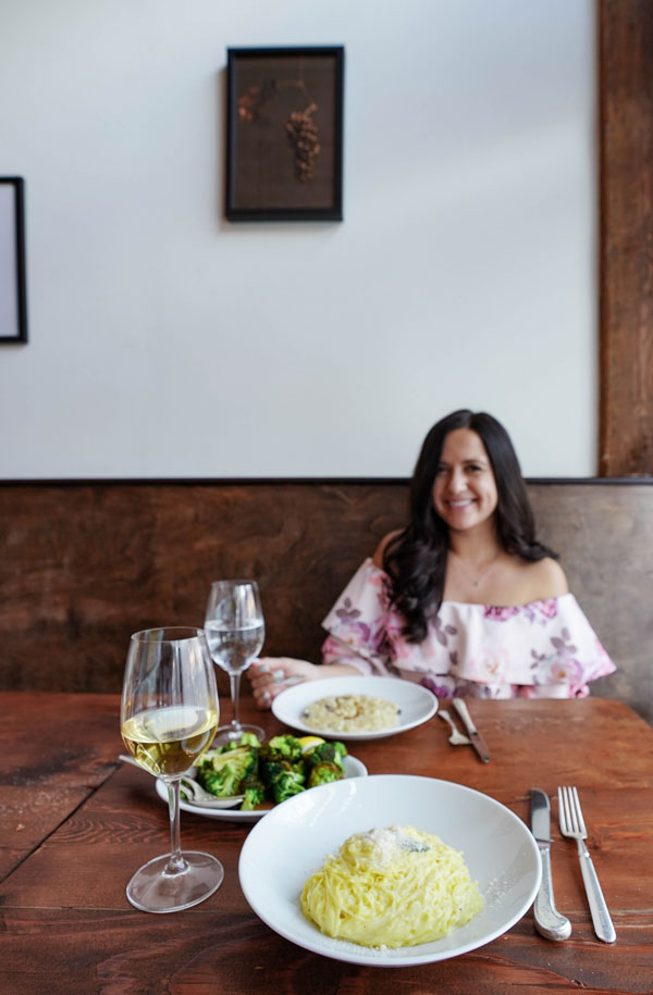 A woman sits at a wooden table in a restaurant, smiling. In front of her are plates of pasta, broccoli, and risotto, along with two glasses of white wine. The background features light walls with minimal decor.