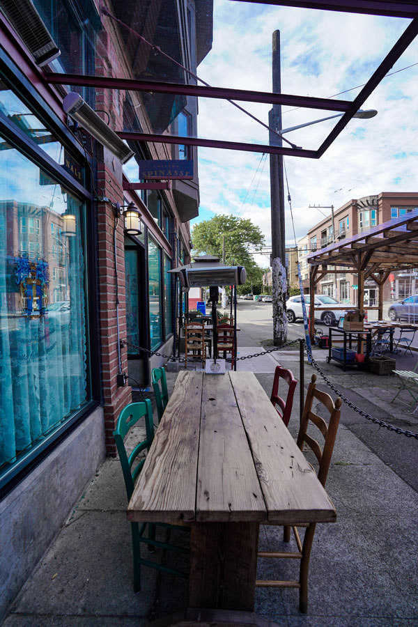Sidewalk café with a rustic wooden table and colorful chairs, set against a city street backdrop. Nearby are storefronts, parked cars, and a covered seating area. A partly cloudy sky completes the scene.