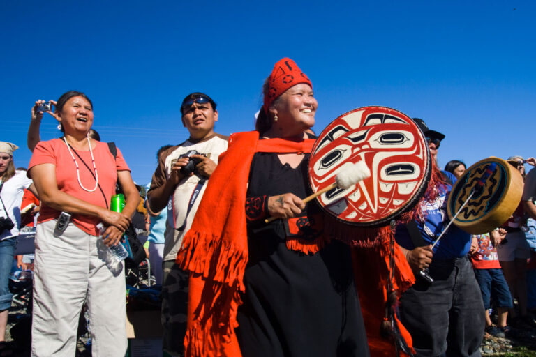 A group of people gathered outdoors in Seattle, with one person in the center wearing traditional attire and holding a decorated drum. Others around are smiling and capturing the moment with cameras. Under the clear blue sky, it's one of those unique things to do in Seattle.