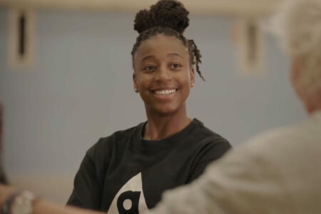 A person with braided hair smiles warmly while talking to someone off-camera. They are wearing a dark t-shirt with a white logo. The background is softly blurred, creating a casual and friendly atmosphere.