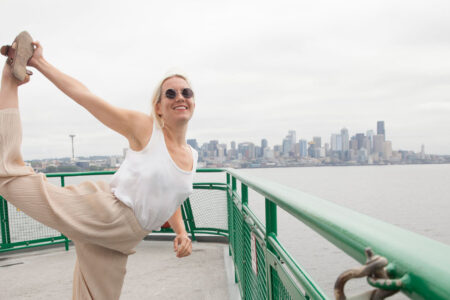 A person wearing sunglasses and a white top performs a standing yoga pose on a ferry deck. The Seattle skyline is visible in the background under a cloudy sky.